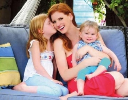 Sarah Rafferty holding her younger daughter, Iris Friday on her lap and being kissed in the cheek by her older daughter, Oona Gray.