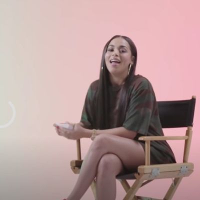 Lauren London is sitting in a chair talking to the camera with pink background.