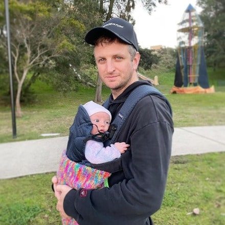 Aaron Glenane posing with his daughter by wearing black cap and black hoodie.