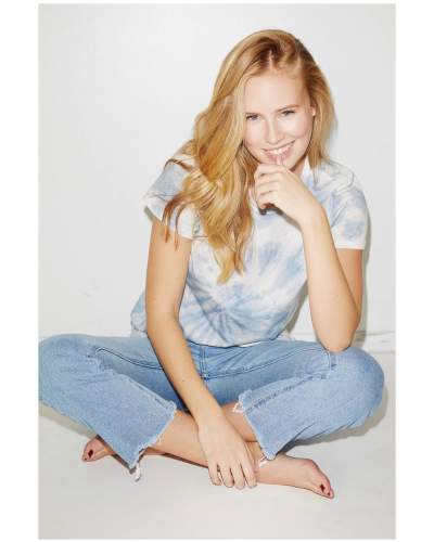 Danika Yarosh posing for the photo with a dark grey hair and a little smile in her face and sitting in the ground with a one hand in her mouth