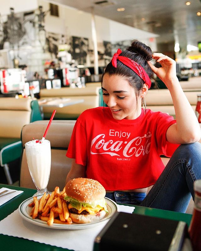 Indiana Massara posing for the photo wearing red tshirt sponsred by Coca-Cola and burger in front of her table.