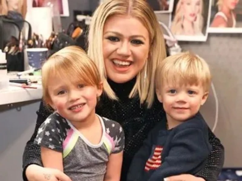 Remington Alexander Blackstock sitting a lap of his mother Kelly Clarkson with his sister River Rose Blackstock posing for the photo wearing hat and in a smiley face