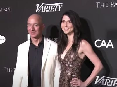 Jeff Bezos is wearing a white suit and a black shirt and Mackenzie Scott is wearing a dress with a lot of patterns on it. 
