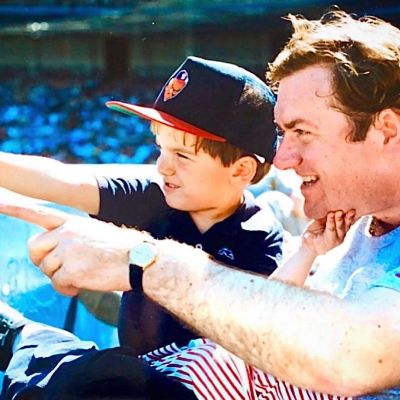 Tim Russert with his son Luke wearing a cap while watching a baseball game.